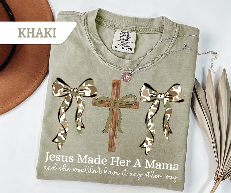 May include: Khaki-colored t-shirt with a cross and two camo bows. The text "Jesus Made Her A Mama and she wouldn't have it any other way" is printed on the shirt. A silver necklace with a pendant is on the shirt.