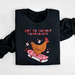 K&ouml;nnte beinhalten: Schwarzes Sweatshirt mit dem Text "OFF TO COMMIT TOMFOOLERY". Ein Cartoon-Huhn mit Zylinder f&auml;hrt auf einem rosa Skateboard mit Herzdekorationen. Das Design beinhaltet Herzen und Liebesbriefe.