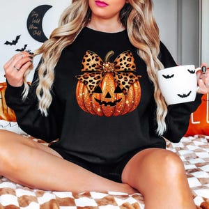 Peut inclure: Sweat-shirt noir avec une citrouille orange scintillante orn&eacute;e d'un n&oelig;ud en imprim&eacute; l&eacute;opard. La citrouille a un visage de citrouille.