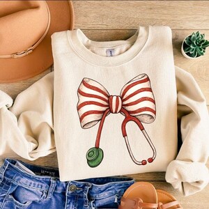 May include: Cream-colored sweatshirt featuring a large bow with red and white stripes and a stethoscope design. The stethoscope has red tubing and a green earpiece. The sweatshirt is paired with blue denim jeans, a brown hat, and sandals.