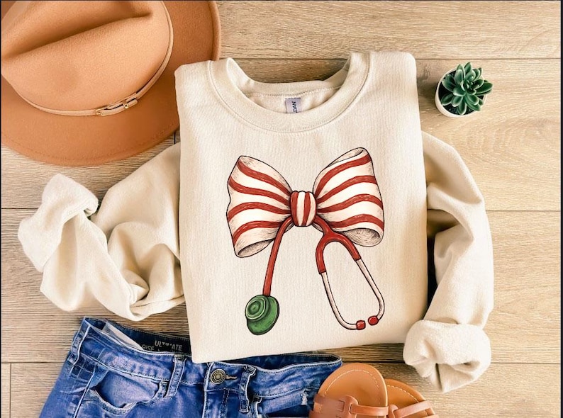May include: Cream-colored sweatshirt featuring a large bow with red and white stripes and a stethoscope design. The stethoscope has red tubing and a green earpiece. The sweatshirt is paired with blue denim jeans, a brown hat, and sandals.