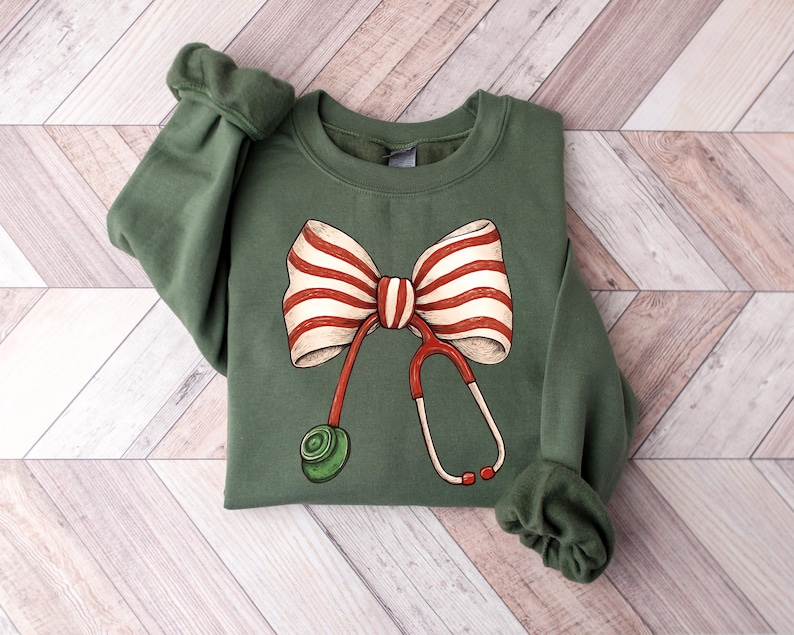 May include: Green sweatshirt featuring a large bow with red and white stripes and a red and white stethoscope design. The sweatshirt has long sleeves with cuffed wrists. The design is centered on the front of the shirt.