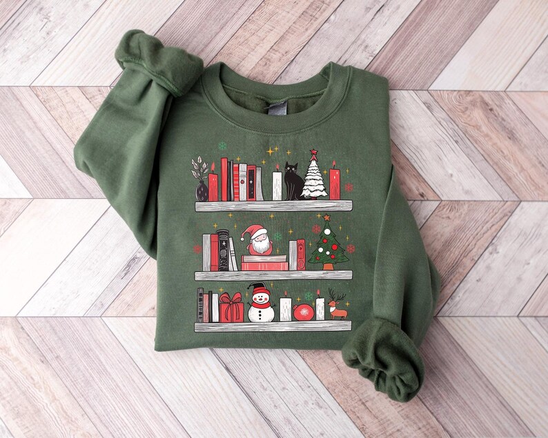 May include: A green sweatshirt with a Christmas-themed design featuring a bookshelf with books, candles, a snowman, Santa Claus, a cat, and a Christmas tree.