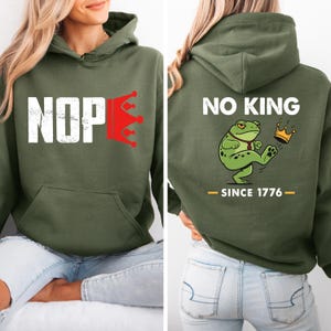 May include: An olive green hoodie with the word "NOPE" in white block letters and a red crown graphic. The back of the hoodie features the text "NO KING" above a cartoon frog kicking a crown, with "SINCE 1776" below.