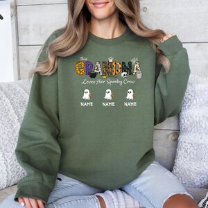 May include: Olive green sweatshirt with the text "This Grandma Loves Her Spooky Crew". The design includes a pumpkin, skull, and ghost graphics. Below the ghosts are three lines labeled "NAME".