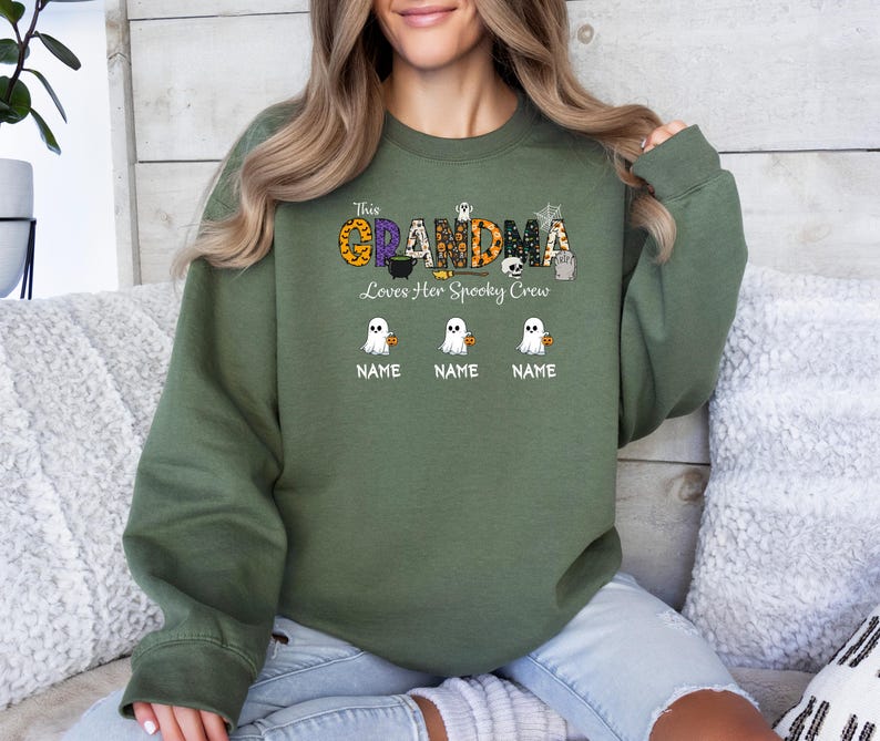May include: Olive green sweatshirt with the text "This Grandma Loves Her Spooky Crew". The design includes a pumpkin, skull, and ghost graphics. Below the ghosts are three lines labeled "NAME".