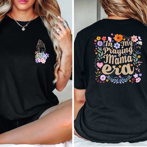 May include: Black t-shirt with a floral design and the text "In my Praying Mama era" on the back. The front of the shirt features a floral design with a praying hand.