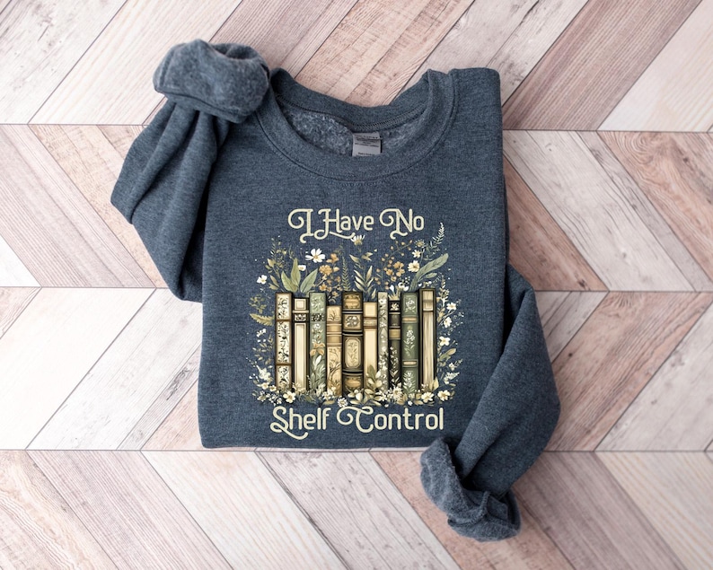 May include: A navy blue sweatshirt with the text "I Have No Shelf Control" above a graphic of vintage books surrounded by flowers and foliage. The sweatshirt is folded on a wooden surface.