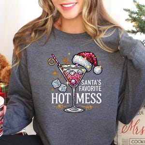 May include: A gray sweatshirt with a sparkly design of a martini glass with a Santa hat on top. The text "Santa's Favorite Hot Mess" is printed below the glass.