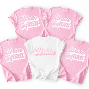 May include: Five t-shirts, four pink and one white, with the words "Bride Squad" and "Bride" in a retro font. The pink shirts have "Bride Squad" in white and pink, while the white shirt has "Bride" in pink.