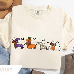 May include: Ivory-colored t-shirt featuring a Halloween-themed dachshund design. The graphic includes dachshunds dressed as a witch, pumpkin, mummy, and ghost, with bats. The shirt has a crew neck and short sleeves. The word "IVORY" is printed at the bottom.