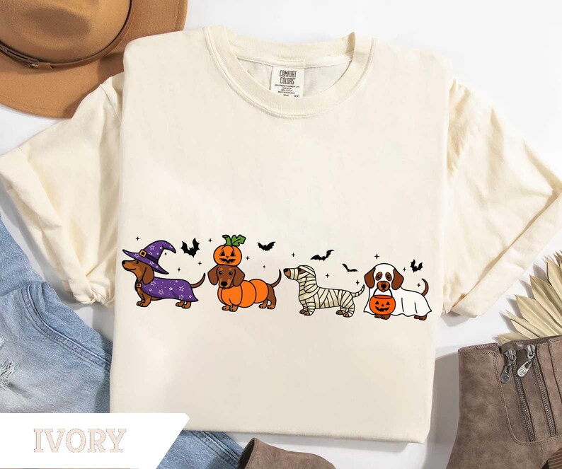 May include: Ivory-colored t-shirt featuring a Halloween-themed dachshund design. The graphic includes dachshunds dressed as a witch, pumpkin, mummy, and ghost, with bats. The shirt has a crew neck and short sleeves. The word "IVORY" is printed at the bottom.