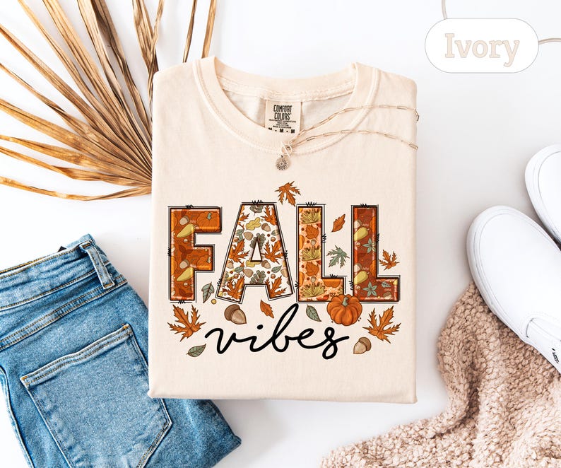 May include: Ivory-colored t-shirt with the words "FALL vibes" in a fall-themed design. The letters "FALL" are filled with autumn leaves, acorns, and pumpkins. The shirt is paired with blue jeans, white shoes, and a brown scarf.