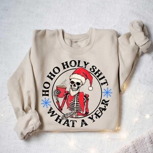 May include: A beige sweatshirt featuring a graphic print of a skeleton wearing a Santa hat and holding a cup of coffee. The graphic is enclosed in a circle with the text "Ho Ho Holy Shit What A Year".