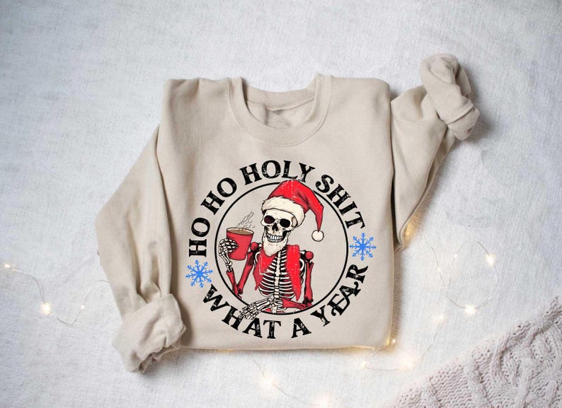 May include: A beige sweatshirt featuring a graphic print of a skeleton wearing a Santa hat and holding a cup of coffee. The graphic is enclosed in a circle with the text "Ho Ho Holy Shit What A Year".
