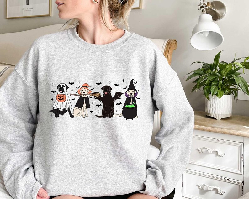May include: Light gray sweatshirt featuring a Halloween-themed graphic. The design includes four dogs dressed in costumes: a ghost, a witch, a bat, and a witch with a cauldron. Black bats are scattered above the dogs.