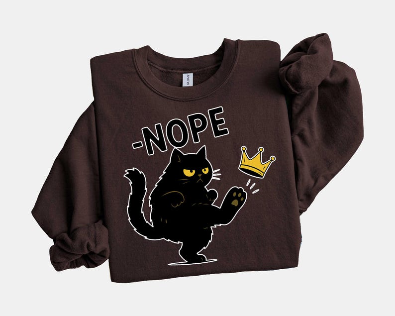May include: A dark brown sweatshirt featuring a black cat kicking a gold crown. The cat has yellow eyes and is outlined in white. The word "-NOPE" is above the cat in white block letters. The sweatshirt is a casual style.