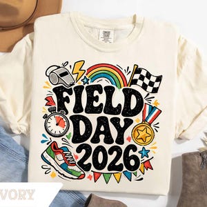 May include: Ivory-colored t-shirt with a colorful graphic that reads "FIELD DAY 2026." The design includes a rainbow, stopwatch, medal, and a checkered flag. A multi-colored running shoe is also featured in the design.