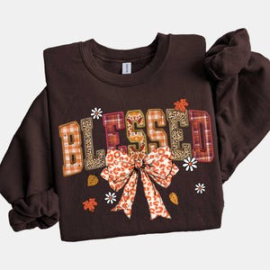 May include: A dark brown sweatshirt with the word "BLESSED" in a patchwork design. The letters feature plaid, leopard print, and orange patterns. A large bow and small flowers and leaves decorate the front.
