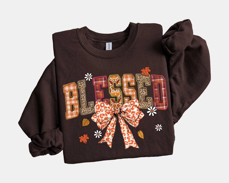 May include: A dark brown sweatshirt with the word "BLESSED" in a patchwork design. The letters feature plaid, leopard print, and orange patterns. A large bow and small flowers and leaves decorate the front.
