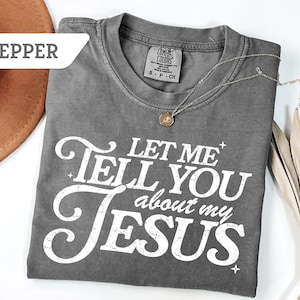 Puede incluir: Una camiseta gris Comfort Colors con el texto "LET ME TELL YOU about my JESUS" en blanco. Un collar plateado con un colgante est&aacute; sobre la camiseta. Un sombrero marr&oacute;n y una planta seca est&aacute;n en el fondo.