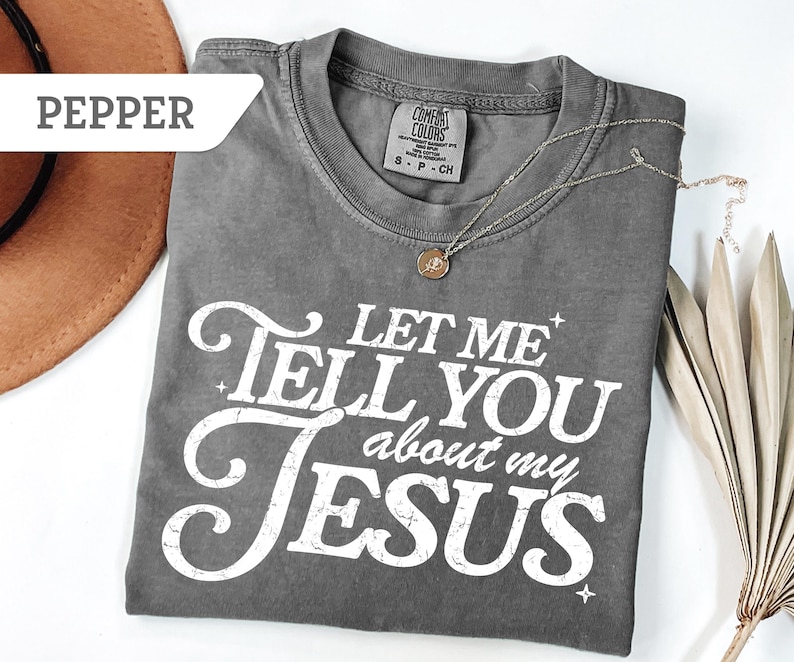 Puede incluir: Una camiseta gris Comfort Colors con el texto "LET ME TELL YOU about my JESUS" en blanco. Un collar plateado con un colgante est&aacute; sobre la camiseta. Un sombrero marr&oacute;n y una planta seca est&aacute;n en el fondo.