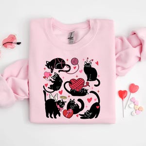 May include: Pink sweatshirt with a black cat design. The cats are wearing angel wings, holding hearts, and playing with yarn. The design is surrounded by hearts and stars.
