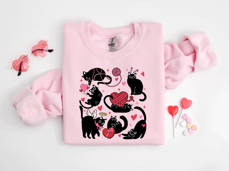 May include: Pink sweatshirt with a black cat design. The cats are wearing angel wings, holding hearts, and playing with yarn. The design is surrounded by hearts and stars.