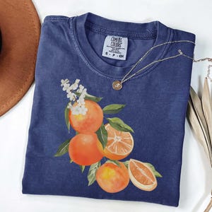 May include: A navy blue t-shirt with a watercolor illustration of oranges and leaves. The shirt has a crew neck and short sleeves. A gold necklace with a small pendant rests on the shirt. The tag reads "COMFORT COLORS".