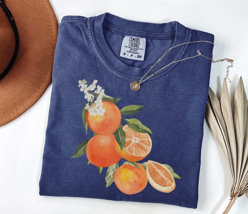May include: A navy blue t-shirt with a watercolor illustration of oranges and leaves. The shirt has a crew neck and short sleeves. A gold necklace with a small pendant rests on the shirt. The tag reads "COMFORT COLORS".