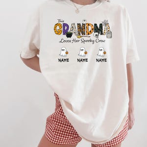 May include: Cream-colored t-shirt with the text "This Grandma Loves Her Spooky Crew." The word "GRANDMA" is in orange, purple, and black patterns. Below are three cartoon ghosts holding pumpkins, with space for names.