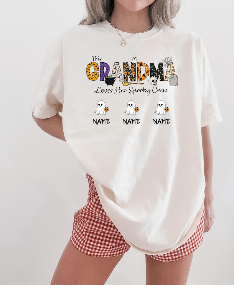 May include: Cream-colored t-shirt with the text "This Grandma Loves Her Spooky Crew." The word "GRANDMA" is in orange, purple, and black patterns. Below are three cartoon ghosts holding pumpkins, with space for names.