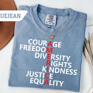May include: Bluejean colored t-shirt with the word "AMERICA" spelled vertically in red, with the words "COURAGE, FREEDOM, DIVERSITY, RIGHTS, KINDNESS, JUSTICE, EQUALITY" in white. A gold necklace with a pendant is draped on the shirt.
