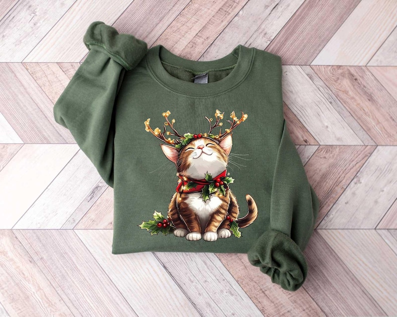 May include: Green Christmas sweater with a brown and white cat wearing reindeer antlers and a red and green holly wreath around its neck.