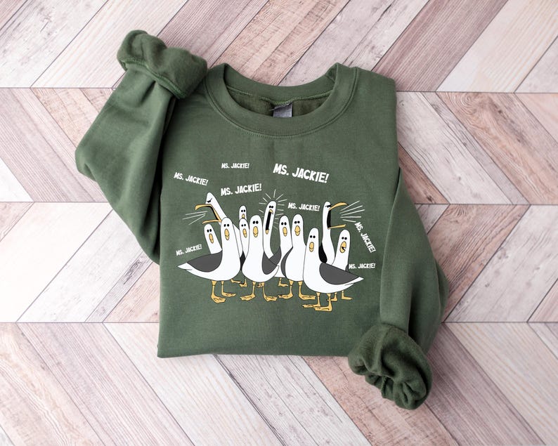 May include: An olive green sweatshirt featuring a cartoon illustration of seagulls with the text "MS. JACKIE!" repeated around the image. The seagulls are in black, white, and yellow.