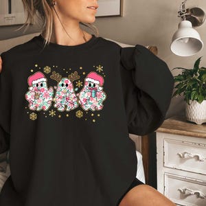 Puede incluir: Sudadera negra con un dise&ntilde;o festivo que presenta tres fantasmas con gorros de Pap&aacute; Noel y patrones florales. Los fantasmas sostienen tazas y est&aacute;n rodeados de copos de nieve y estrellas.