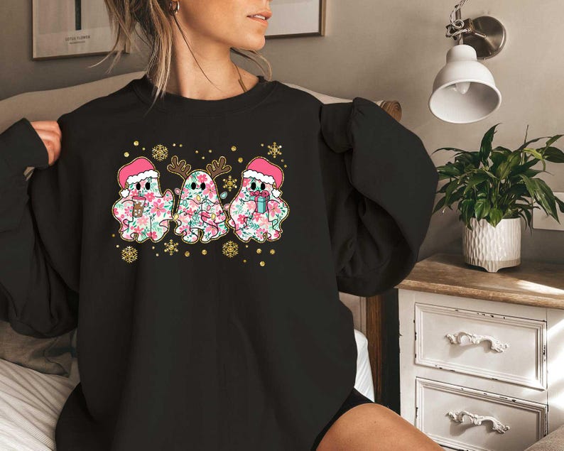 Puede incluir: Sudadera negra con un dise&ntilde;o festivo que presenta tres fantasmas con gorros de Pap&aacute; Noel y patrones florales. Los fantasmas sostienen tazas y est&aacute;n rodeados de copos de nieve y estrellas.