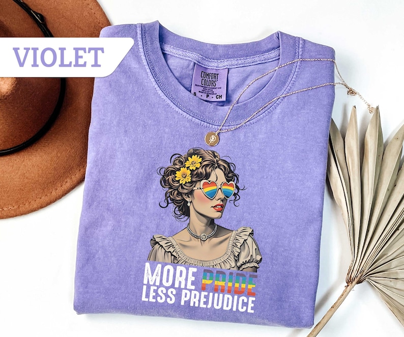May include: Violet Comfort Colors t-shirt featuring a graphic of a woman wearing rainbow heart-shaped sunglasses and flowers in her hair. The text "MORE PRIDE LESS PREJUDICE" is printed below the graphic. A gold necklace is draped on the shirt.