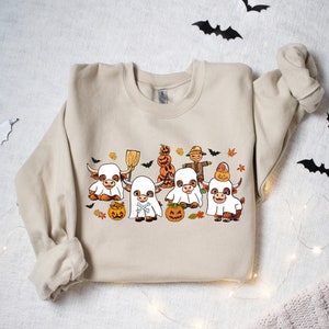 Op de afbeelding: Een beige sweatshirt met een Halloween-thema ontwerp met vier bruine en witte koeien die verkleed zijn als geesten. De koeien houden pompoenen en een bezem vast. Er zijn ook vleermuizen en bladeren in het ontwerp.