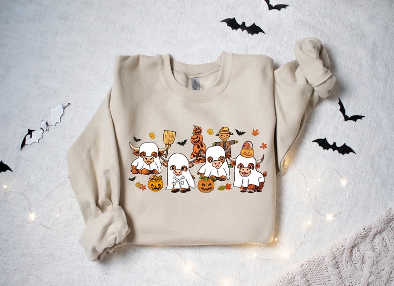 Op de afbeelding: Een beige sweatshirt met een Halloween-thema ontwerp met vier bruine en witte koeien die verkleed zijn als geesten. De koeien houden pompoenen en een bezem vast. Er zijn ook vleermuizen en bladeren in het ontwerp.