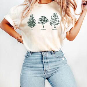 Pu&ograve; includere: Una maglietta bianca con una grafica di tre alberi, etichettati "Fir", "Dogwood" e "Tamarack". La modella indossa la maglietta con jeans blu.