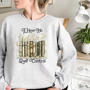 May include: A light gray sweatshirt featuring the phrase "I Have No Shelf Control" in elegant script. The design includes a stack of vintage-style books with floral accents. The sweatshirt is a comfortable, casual garment.