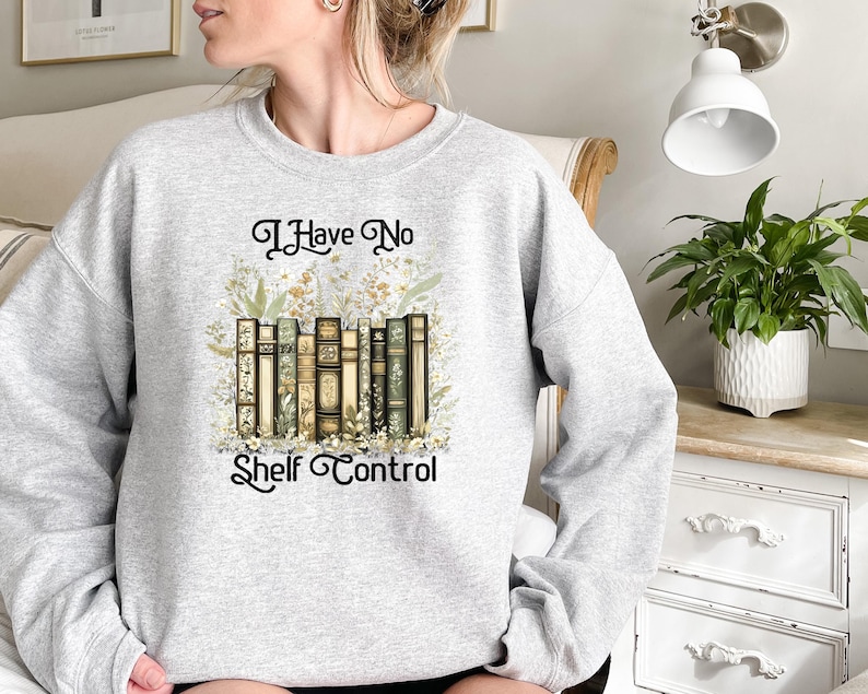May include: A light gray sweatshirt featuring the phrase "I Have No Shelf Control" in elegant script. The design includes a stack of vintage-style books with floral accents. The sweatshirt is a comfortable, casual garment.