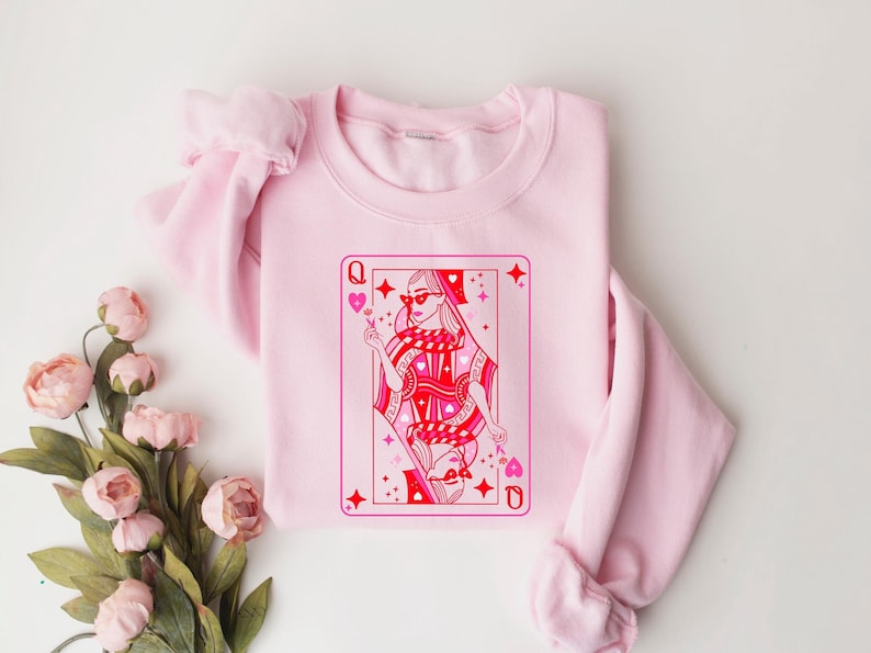 May include: Pink sweatshirt with a graphic of a queen of hearts playing card. The queen is wearing sunglasses and a crown. The card is outlined in red and has a pink background.
