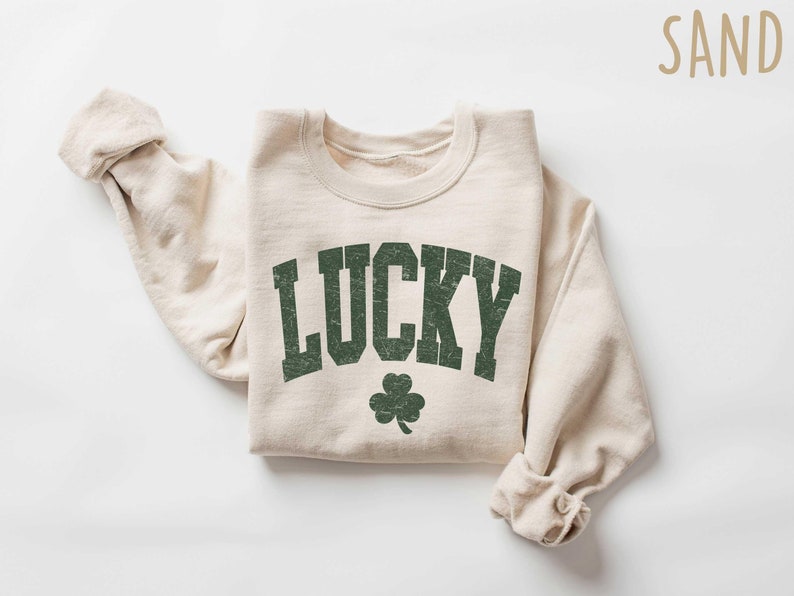 Peut inclure: Un sweat-shirt couleur cr&egrave;me avec le mot "LUCKY" imprim&eacute; en vert avec un tr&egrave;fle vert en dessous. Le sweat-shirt est pli&eacute; et pos&eacute; &agrave; plat sur une surface blanche.
