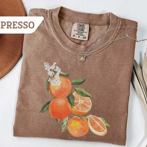 May include: Espresso-colored t-shirt with a watercolor illustration of oranges and white flowers. The shirt has a crew neck and short sleeves. A silver necklace with a small pendant is draped across the shirt.