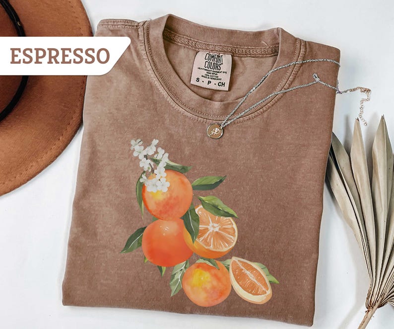 May include: Espresso-colored t-shirt with a watercolor illustration of oranges and white flowers. The shirt has a crew neck and short sleeves. A silver necklace with a small pendant is draped across the shirt.