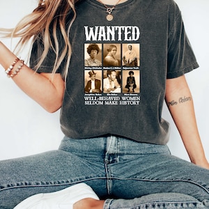 K&ouml;nnte beinhalten: Ein dunkelgraues T-Shirt mit einer Grafik im Vintage-Stil, die Portr&auml;ts einflussreicher Frauen zeigt. Die Grafik enth&auml;lt den Text "WANTED" und "WELL-BEHAVED WOMEN SELDOM MAKE HISTORY".
