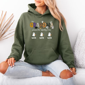 May include: An olive green hooded sweatshirt with the text "This Grandma Loves Her Spooky Crew" in a Halloween-themed design. The design includes ghosts and pumpkins. Below the text are three ghost graphics with space for names.