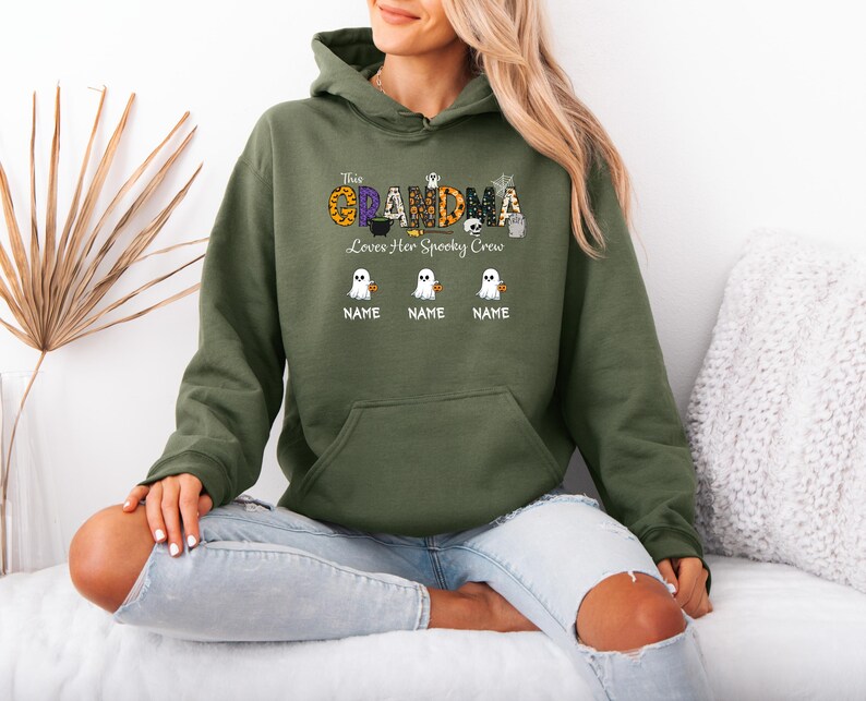 May include: An olive green hooded sweatshirt with the text "This Grandma Loves Her Spooky Crew" in a Halloween-themed design. The design includes ghosts and pumpkins. Below the text are three ghost graphics with space for names.